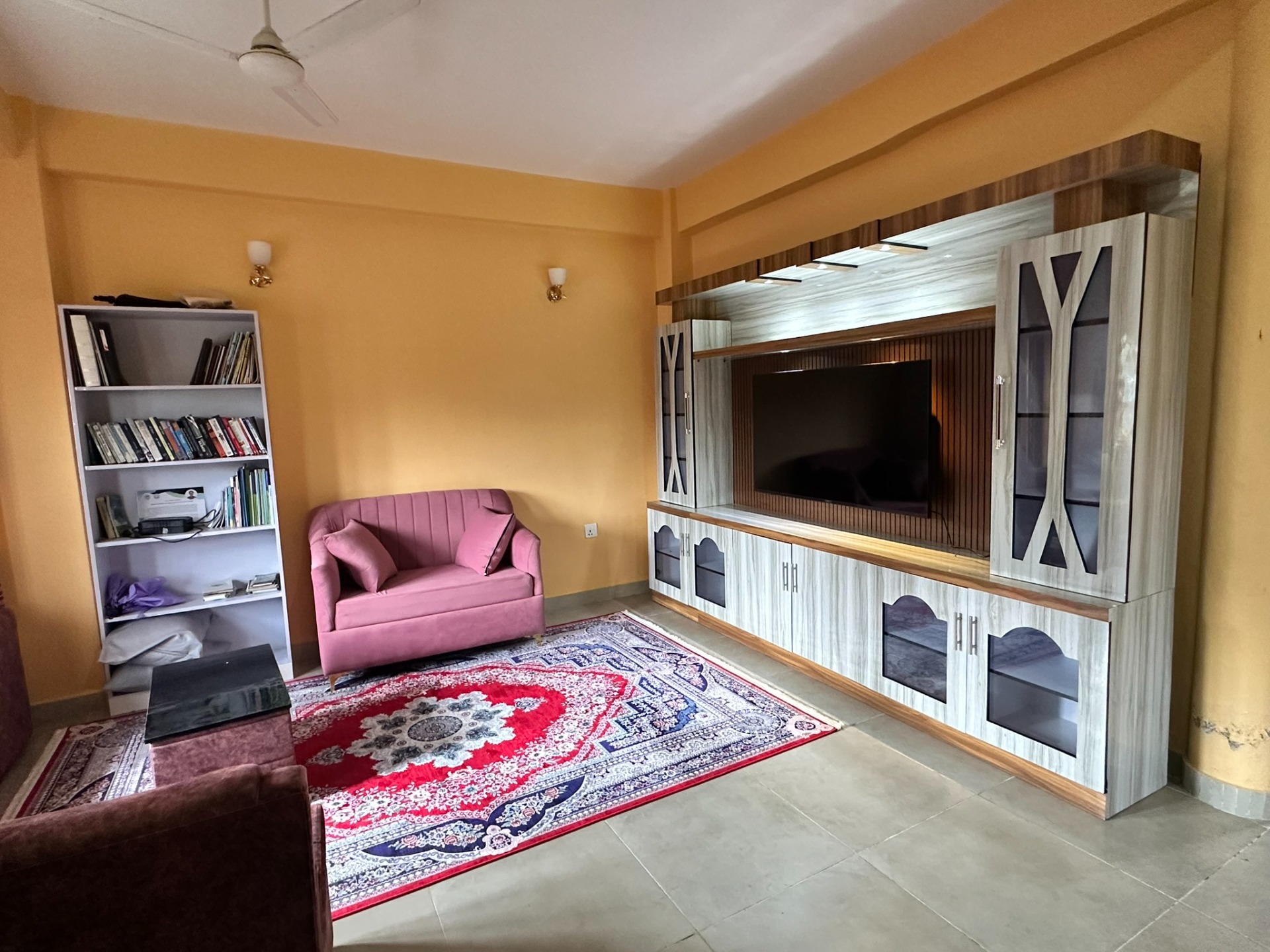 Woonkamer met roze bank, kleurrijk tapijt, boekenkast en groot tv-meubel met open haard.