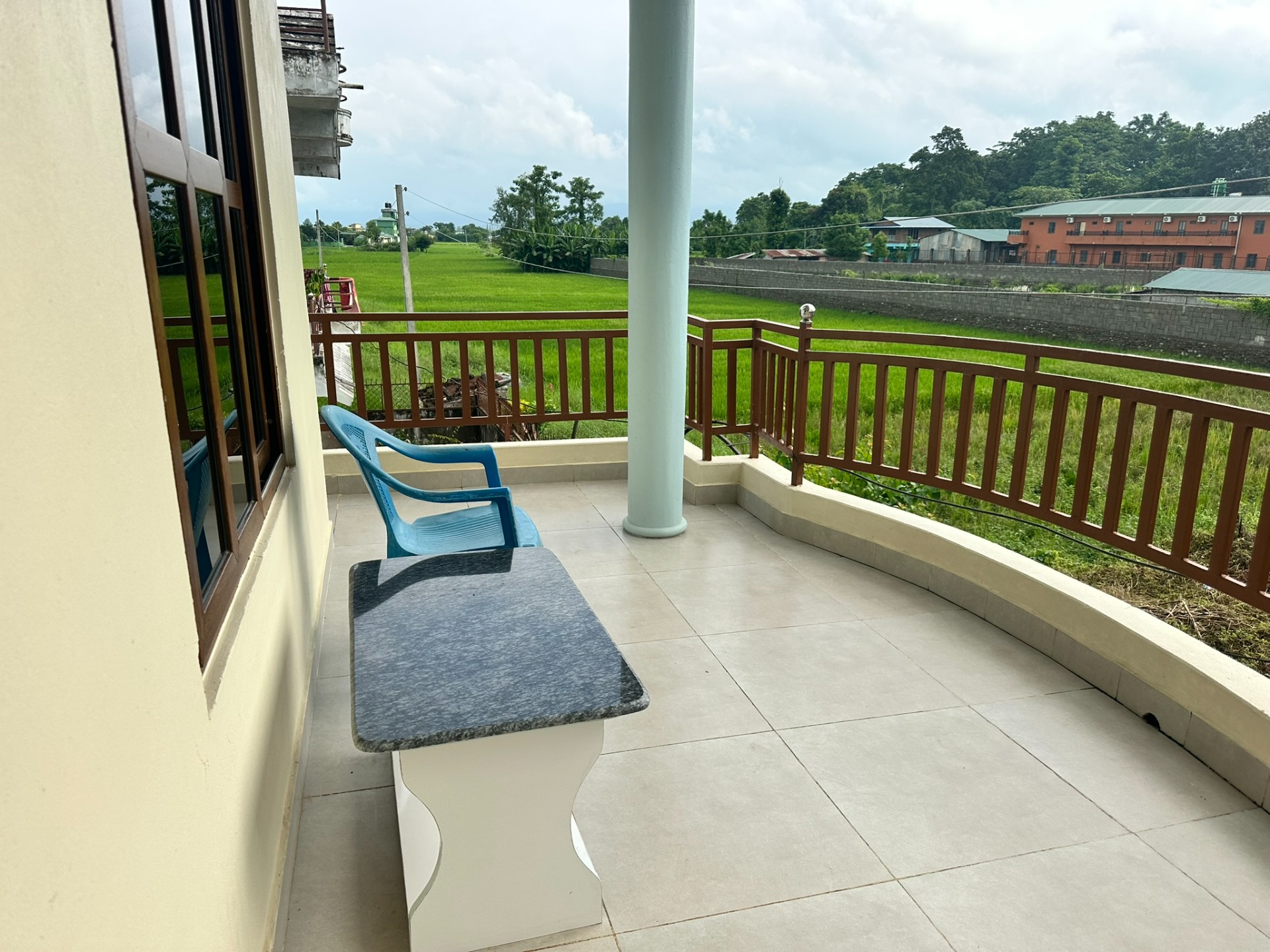 Terras met blauwe stoel en marmeren tafel, uitzicht op groene velden en gebouwen onder bewolkte lucht.