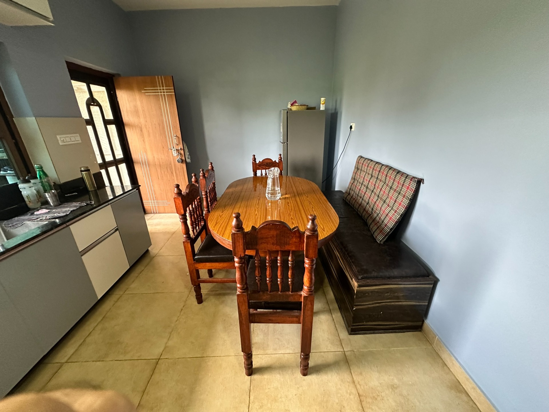 Houten eettafel met stoelen, glazen pot en keukenhoek met deur en bank in lichte woonkamer.